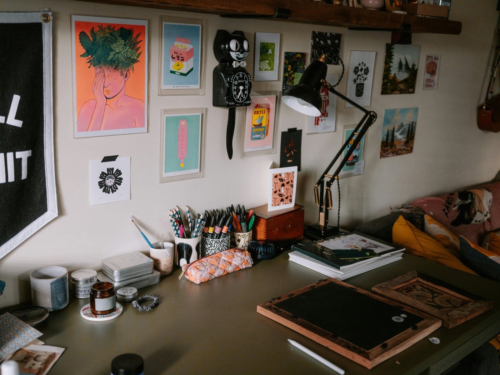 A creative's desk under a wall of colorful prints, with a lamp, pencil cups and sketchbooks