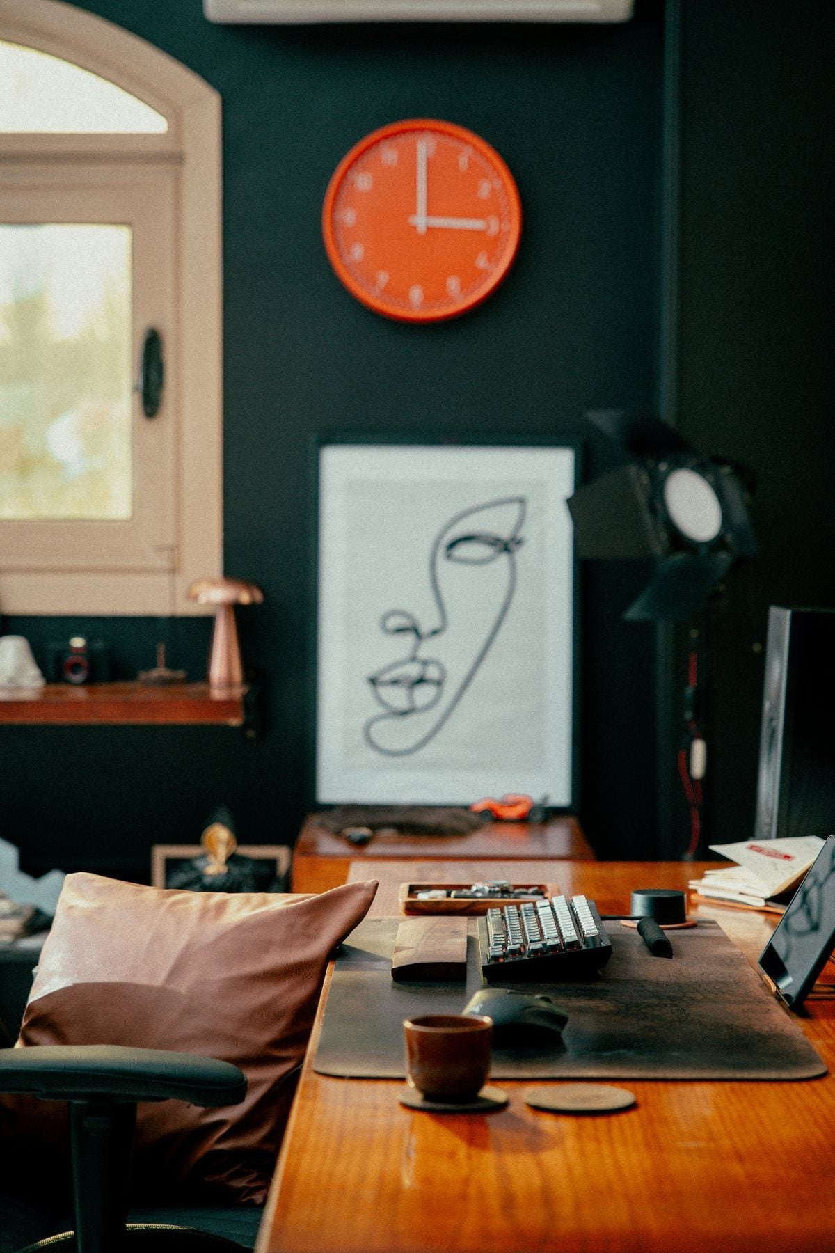 A desk against a dark green wall with an orange clock, a line drawing and a warm wooden desktop
