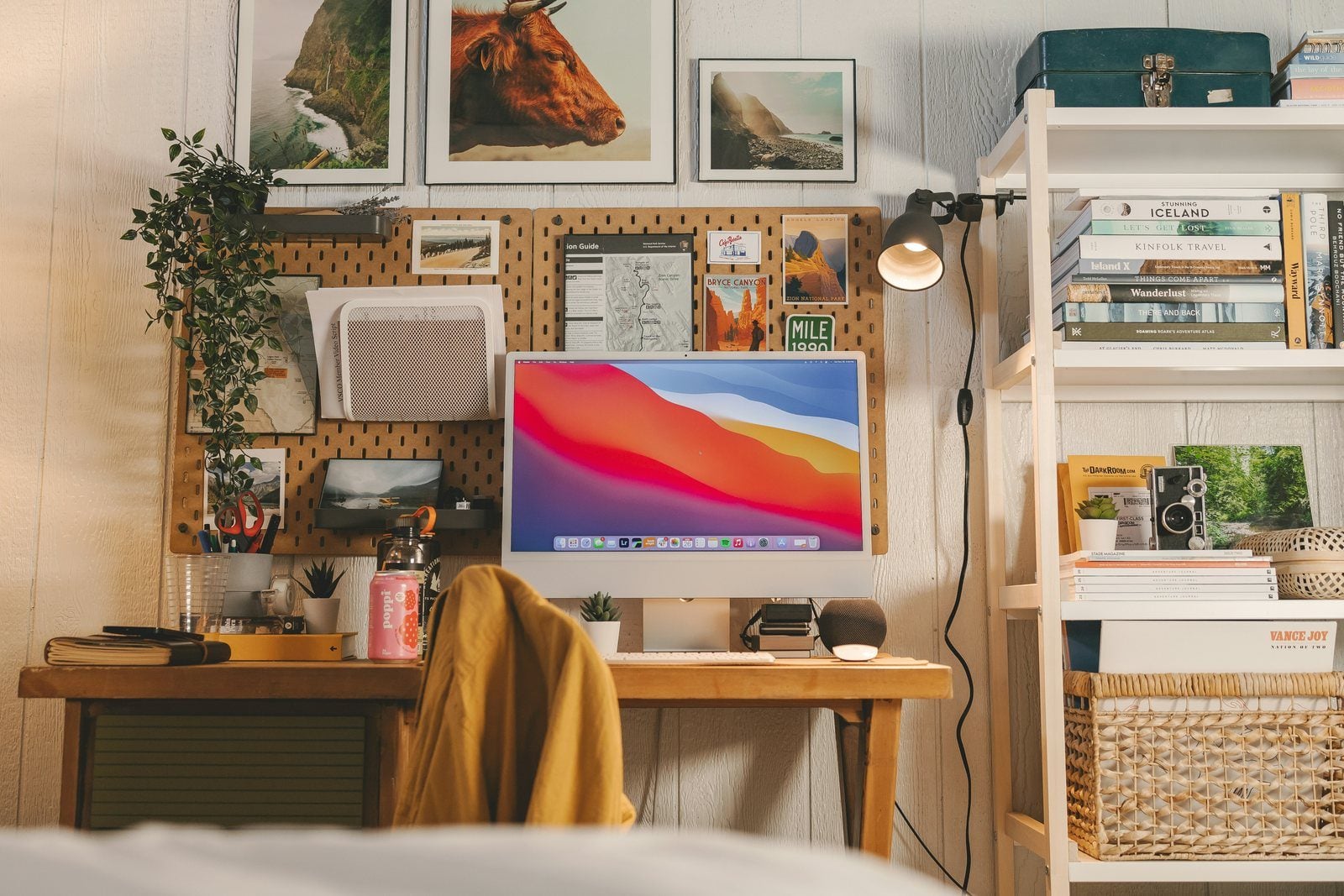 A home studio desk with a pegboard, framed prints, shelves of books and a colorful screen
